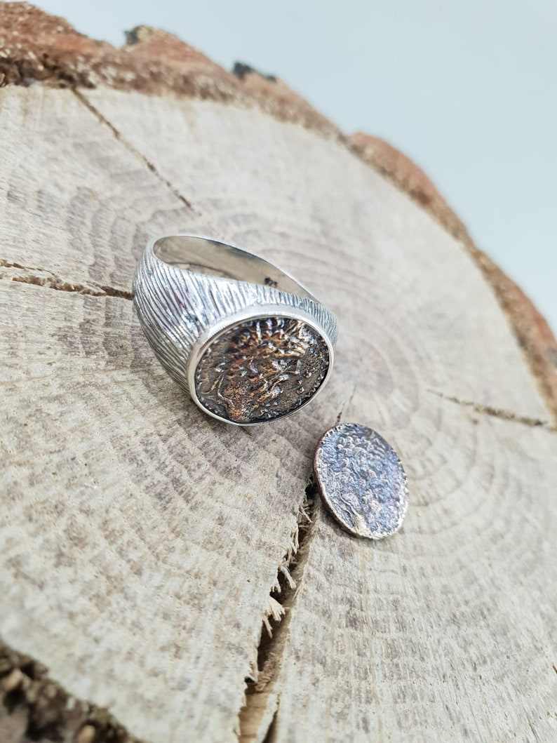 Puede incluir: Un anillo de plata con una banda texturizada y un dise&ntilde;o de moneda redonda de plata oxidada. La moneda presenta un perfil detallado de la cara de una persona.