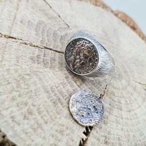 Puede incluir: Un anillo de plata con una banda texturizada y un dise&ntilde;o redondo y detallado similar a una moneda que presenta una cara. La moneda est&aacute; separada del anillo y se encuentra sobre una superficie de madera.