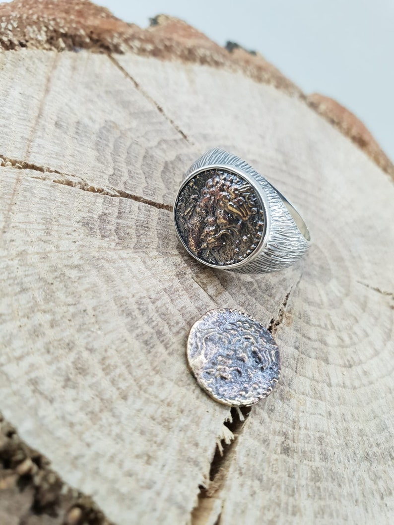 Puede incluir: Un anillo de plata con una banda texturizada y un dise&ntilde;o redondo y detallado similar a una moneda que presenta una cara. La moneda est&aacute; separada del anillo y se encuentra sobre una superficie de madera.