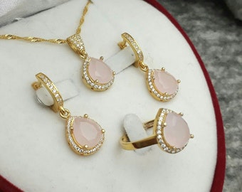 May include: A gold jewelry set with a necklace, earrings, and a ring. Each piece features a pink stone surrounded by a halo of clear stones.