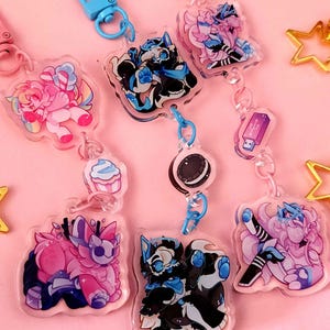 May include: A set of seven colorful keychains featuring cartoon characters and designs. The keychains are made of clear acrylic and have a variety of colors, including pink, blue, black, white, and rainbow. The keychains are attached to silver chains and have a silver clasp.