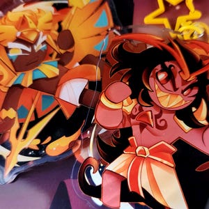 May include: Two cartoon character acrylic charms. The first charm is a red and orange character with a red and yellow crown and a red and yellow cape. The second charm is a red and orange character with a red and yellow crown and a red and yellow cape. The charms are on a black background.
