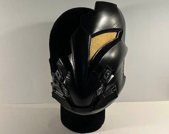 Könnte beinhalten: Eine schwarze Plastikmaske mit einem goldfarbenen Visier und mehreren kleinen Details. Die Maske steht auf einem schwarzen Ständer.