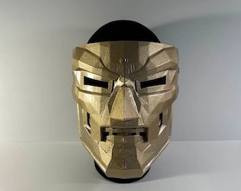 May include: A gold-colored plastic mask with a geometric design. The mask has two eye holes and a mouth opening.