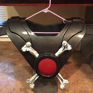 May include: A black and silver costume piece with a red circle and two white bones crossed over it.