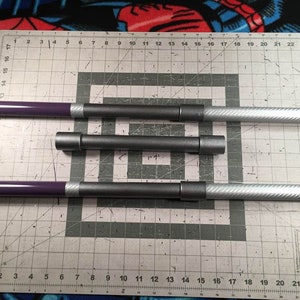May include: Three metal rods with different lengths. The rods are painted purple and gray with a silver carbon fiber pattern. The rods are laid out on a gray cutting mat with white grid lines.