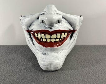 May include: A white, half-face mask with a sinister grin. The mask features a wide, red-painted mouth with rows of yellow teeth. Black accents add depth to the white surface, creating a distressed look. The nose is also white, and the mask is designed to cover the lower half of the face.