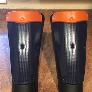 Può includere: Un paio di parastinchi blu navy e neri con dettagli arancioni. I parastinchi hanno una superficie strutturata e sono fissati con rivetti. La parte superiore di ogni protezione è arancione e la parte inferiore è nera.