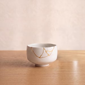 Könnte beinhalten: Eine kleine weiße Keramikschale mit einer Kintsugi-Reparatur, bei der goldfarbene Adern über die Außenfläche verlaufen. Die Schale hat eine abgerundete Form mit einem kurzen Fuß und eine glatte, glänzende Oberfläche auf dem weißen Material.