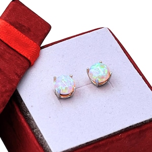 May include: A pair of opal stud earrings displayed in an open red velvet gift box with a red ribbon. The earrings have a silver setting and a round, iridescent opal stone. The box has a white interior.