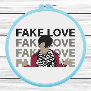 May include: A cross-stitch embroidery hoop with a white background and a black and white image of a person with black hair wearing a red jacket and a black and white striped shirt. The text "FAKE LOVE" is repeated four times in black text.