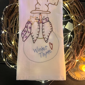 May include: White hand towel with an embroidered snowman design. The snowman has a brown hat, carrot nose, and a purple scarf. The text "Winter Thyme" is embroidered below the snowman, with a small heart symbol.