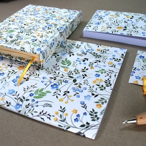 Puede incluir: Un conjunto de artículos de papelería con un patrón floral. El conjunto incluye una libreta, un bloc de notas, un marcapáginas y un bolígrafo. La libreta y el bloc de notas tienen un fondo blanco con flores azules y amarillas. El marcapáginas tiene un fondo blanco con flores azules y amarillas y una cinta amarilla. El bolígrafo es marrón con un cuerpo de madera.
