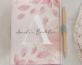 May include: A personalized notebook with a pink floral design and the name "Amelie Bachler" in script. The cover features a large white letter "A" and scattered pink leaves. A wooden pen with a gray elastic band is next to the notebook.