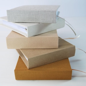 Könnte beinhalten: Ein Stapel von fünf Hardcover-Büchern in verschiedenen neutralen Farbtönen. Die Bücher haben strukturierte Einbände in Grau-, Beige- und Brauntönen. Jedes Buch hat ein Leseband. Die Bücher sind übereinander gestapelt.