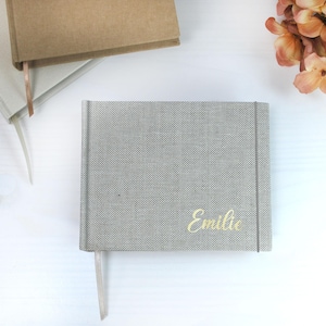 Könnte beinhalten: Ein graues, mit Stoff bezogenes Notizbuch mit dem Namen "Emilie" in goldener Schrift. Ein helles Band-Lesezeichen ragt aus dem Notizbuch heraus. Weitere Notizbücher sind im Hintergrund zu sehen, zusammen mit einem Blumenarrangement.