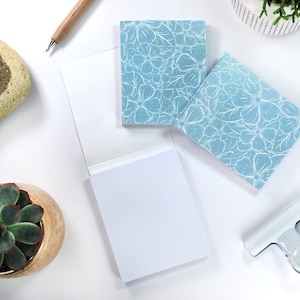 Könnte beinhalten: Zwei blaue Notizblöcke mit weißen Blumenmustern, eine weiße Klammer und zwei Topfpflanzen auf einer weißen Oberfläche.