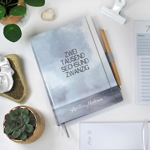 Peut inclure: Un carnet à couverture rigide avec le texte "ZWEI TAUSEND SECHSUND ZWANZIG" en noir. La couverture présente un motif aquarelle dans les tons gris et bleu. Un stylo en bois avec une bande grise repose sur le carnet. Une petite plante succulente dans un pot doré est également visible.