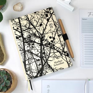 May include: A cream-coloured journal with a black tree branch design, the text "2025" and "Markus Schreiber". A wooden pen with a black band, a "To Do" notepad, and small potted plants are also visible.