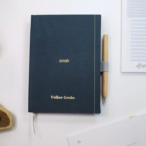 May include: A dark teal 2026 planner with gold lettering, a matching pen, and a notepad. The planner has the name "Volker Grobe" printed on the cover. A light grey pen holder is attached to the planner.