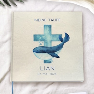 Puede incluir: Libro de visitas de bautizo color crema con una ilustración de acuarela de una ballena azul y una cruz. El texto "MEINE TAUFE" está en la parte superior, con el nombre "LIAN" y la fecha "02. MAI 2026" debajo.