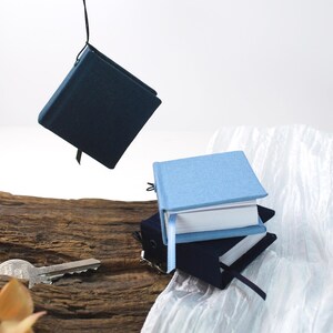 Könnte beinhalten: Mehrere kleine, quadratische Notizbücher mit blauen Einbänden und weißen Seiten. Eines ist an einer schwarzen Schnur aufgehängt. Ein Schlüssel liegt auf einem Holzstück. Jedes Notizbuch hat ein Leseband. Die Notizbücher sind gestapelt.