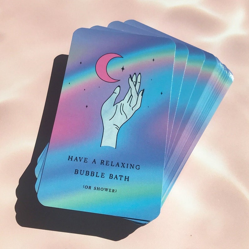 May include: A deck of oracle cards with a pastel rainbow design. Each card features a hand reaching for a pink crescent moon. The text on the card reads "HAVE A RELAXING BUBBLE BATH (OR SHOWER)".