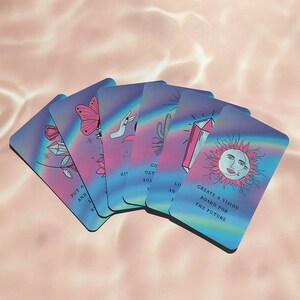 May include: A deck of oracle cards with a pastel rainbow design. Each card features a unique illustration, including a butterfly, rose, and sun with text that reads "Create a vision board for the future."