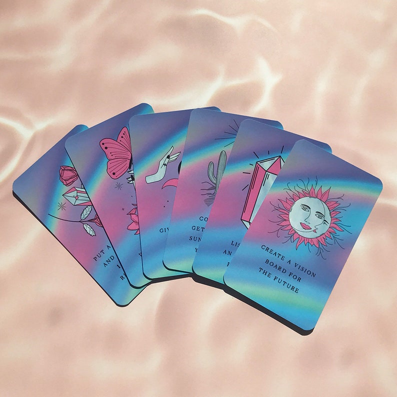 May include: A deck of oracle cards with a pastel rainbow design. Each card features a unique illustration, including a butterfly, rose, and sun with text that reads "Create a vision board for the future."