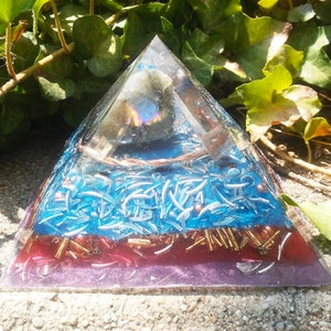 May include: A clear glass pyramid with a layered design. The pyramid has a blue layer with silver flakes, a red layer with gold flakes, and a purple base. A clear crystal is visible in the center of the pyramid.