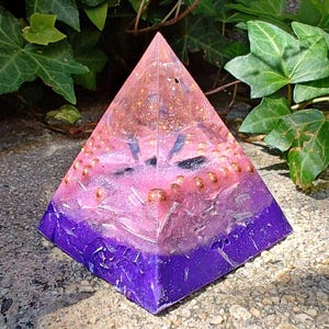 May include: A pyramid-shaped resin object featuring a gradient color scheme that transitions from a deep purple base to a translucent pink top. The interior contains embedded gold-colored metallic spheres and scattered silver-toned flakes.