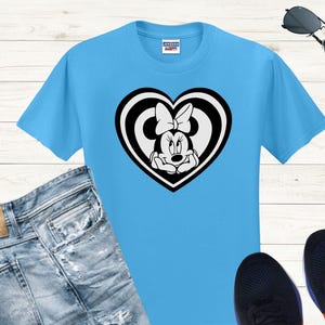May include: A light blue t-shirt featuring a black and white Minnie Mouse graphic inside a heart shape. The graphic shows Minnie Mouse with her signature bow. The shirt is paired with blue jeans, black sneakers, and sunglasses.