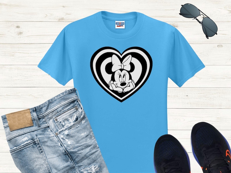May include: A light blue t-shirt featuring a black and white Minnie Mouse graphic inside a heart shape. The graphic shows Minnie Mouse with her signature bow. The shirt is paired with blue jeans, black sneakers, and sunglasses.