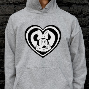 May include: A gray hoodie featuring a black and white Minnie Mouse design inside a heart shape. The Minnie Mouse graphic includes her face and bow. The hoodie has a front pocket and a hood.