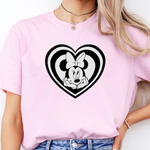 May include: A light pink t-shirt featuring a black and white Minnie Mouse design inside a heart shape. The Minnie Mouse graphic includes her face, bow, and hands. The shirt is made of a soft material.