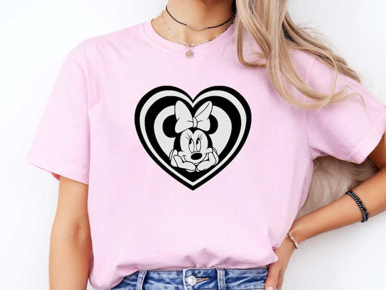 May include: A light pink t-shirt featuring a black and white Minnie Mouse design inside a heart shape. The Minnie Mouse graphic includes her face, bow, and hands. The shirt is made of a soft material.