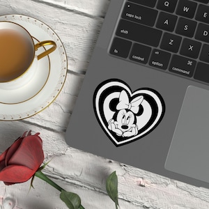 May include: A black and white Minnie Mouse sticker in a heart shape is on a laptop. A cup of tea in a white teacup with a gold rim sits on a saucer. A red rose is on the table.