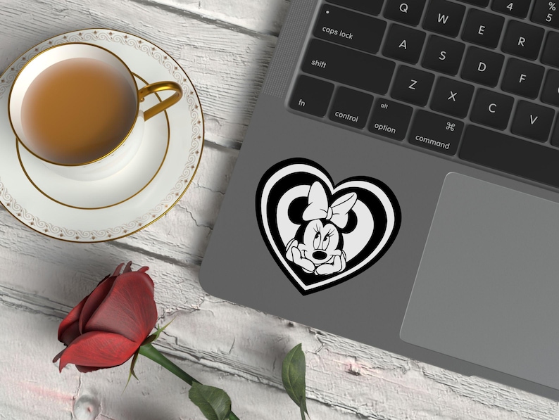 May include: A black and white Minnie Mouse sticker in a heart shape is on a laptop. A cup of tea in a white teacup with a gold rim sits on a saucer. A red rose is on the table.