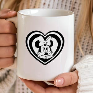 May include: White ceramic mug featuring a black and white Minnie Mouse design inside a heart shape. The design includes Minnie's face and bow. The mug has a curved handle and is held by a person.