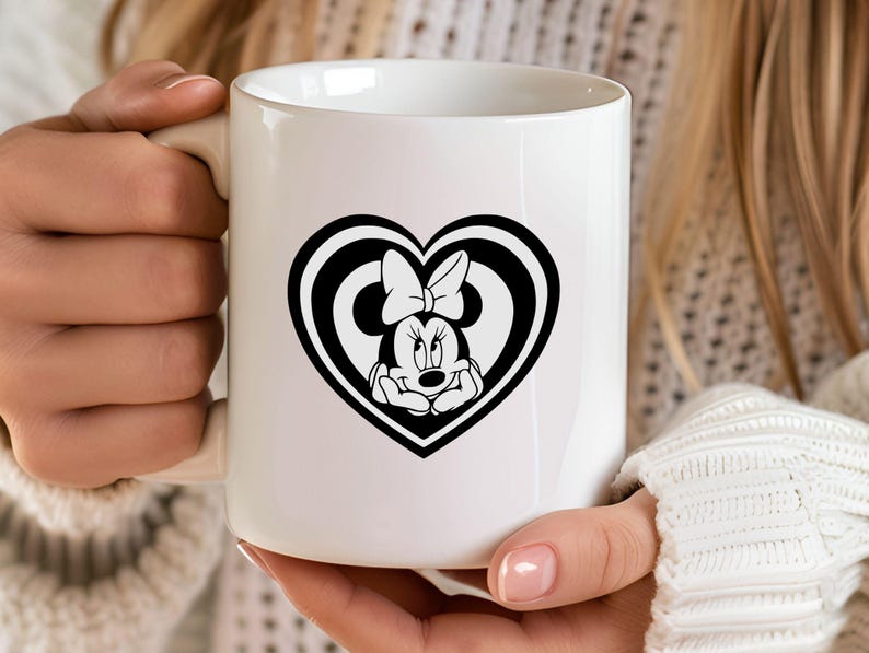 May include: White ceramic mug featuring a black and white Minnie Mouse design inside a heart shape. The design includes Minnie's face and bow. The mug has a curved handle and is held by a person.