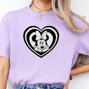 May include: A light purple t-shirt featuring a black and white heart-shaped design with Minnie Mouse. The graphic shows Minnie Mouse inside a heart with a bow. The t-shirt is made of a soft material.
