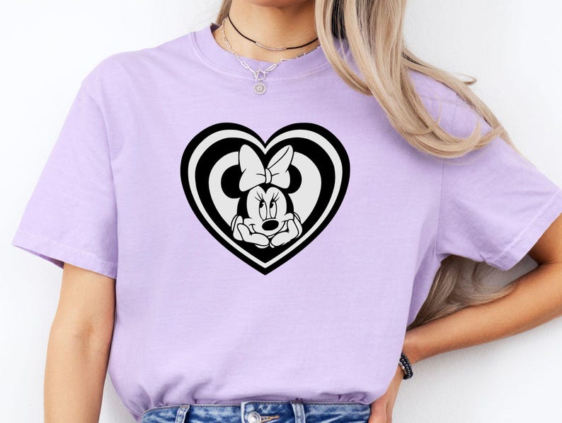 May include: A light purple t-shirt featuring a black and white heart-shaped design with Minnie Mouse. The graphic shows Minnie Mouse inside a heart with a bow. The t-shirt is made of a soft material.