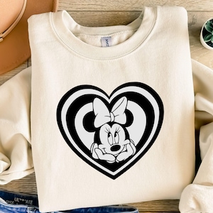 May include: Cream-colored sweatshirt featuring a black and white Minnie Mouse design inside a heart shape. The Minnie Mouse graphic includes her signature bow. The sweatshirt is made of a soft material and is suitable for casual wear.