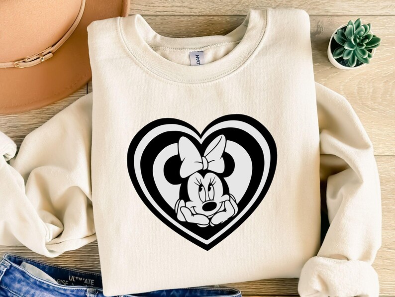 May include: Cream-colored sweatshirt featuring a black and white Minnie Mouse design inside a heart shape. The Minnie Mouse graphic includes her signature bow. The sweatshirt is made of a soft material and is suitable for casual wear.
