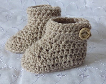 May include: A pair of handmade, beige crochet baby booties. Each bootie features a rolled cuff and a wooden button with the words "handmade with love" engraved on it. The booties are made with a thick yarn.