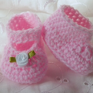 May include: A pair of pink crocheted baby booties. Each bootie is adorned with a white rose and a green ribbon. The booties have a strap and are made with a delicate, openwork stitch.