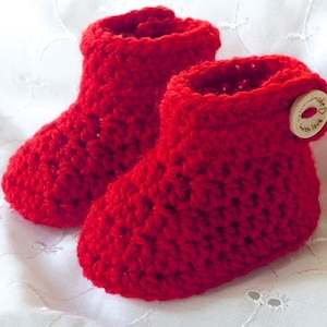 Può includere: Un paio di scarpine da neonato all'uncinetto, fatte a mano, di colore rosso vivo. Le scarpine sono realizzate con un filato spesso, con una chiusura a bottone sul lato di una scarpina. Il bottone è di colore legno chiaro e riporta la scritta "handmade with love".