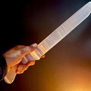 May include: A translucent, off-white selenite crystal carved into the shape of a knife. The object features a smooth, elongated blade with a distinct handle section, displaying natural fibrous mineral striations throughout its semi-opaque surface.