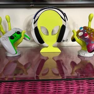 Op de afbeelding: Drie 3D-geprinte handen die een groen-witte videogamecontroller, een kleurrijke videogamecontroller en een zwart-witte headset vasthouden. De handen zijn geel geverfd en houden de controllers en de headset rechtop.