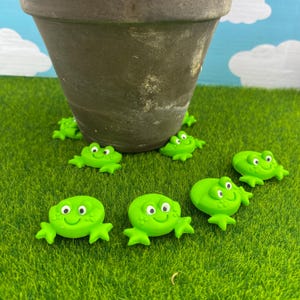 May include: Several small, bright green frog decorations with smiling faces are arranged on artificial green grass. A brown flower pot is in the background. The frogs are small, likely less than 5 cm in size.